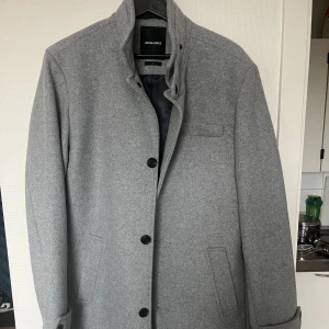 Grå rock från Jack & Jones - Snygg grå rock från Jack & Jones i storlek L. Den har en stilren design med knappar framtill och en hög krage. Perfekt för kyligare dagar med sin klassiska look. Använd en gång. Nypris 1100kr
