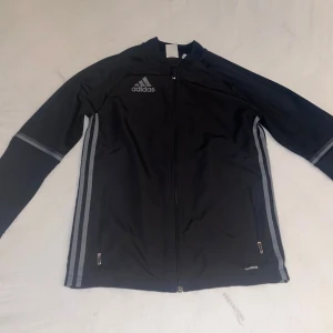 Svart träningsjacka från Adidas - Säljer en svart träningsjacka från Adidas med dragkedja och grå detaljer. Jackan har långa ärmar och är perfekt för träning. Den har också två fickor med dragkedjor framtill.