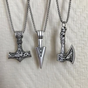 Tre halsband med vikingainspirerade hängen - Säljer tre halsband med vikingainspirerade hängen i silverfärg. Ett hänge är en detaljerad torshammare, ett är en stiliserad pilspets och det tredje är en yxa med intrikata mönster. Kedjorna är i matchande silverfärg och ger en rustik känsla.