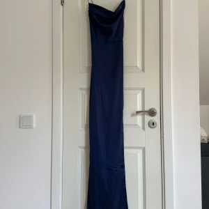 Marinblå långklänning med smala axelband - Elegant marinblå långklänning med smala axelband och draperad urringning. Perfekt för en stilren look. Klänningen har en slät och glansig yta som ger en lyxig känsla. Klänningen är fodrad och har även en slits💕