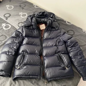 Mörk blå dunjacka från Moncler - Säljer en snygg svart dunjacka från Moncler med huva och dragkedja. Jackan har en quiltad design och praktiska fickor med knappar. Insidan är brun och ger en fin kontrast. Perfekt för kyliga dagar!