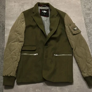 Olivgrön jacka/kofta från Dry Laundry Japan - Säljer en olivgrön jacka från Dry Laundry Japan med quiltade beige ärmar och dragkedjor på fickorna. Jackan har en stilren design med en klassisk krage och knappar framtill. Perfekt för höst och vår!