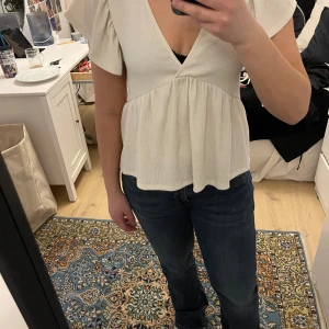 Vit blus från Gina Tricot - Säljer en söt vit blus från Gina Tricot med v-ringning och volangdetaljer. Blusen har korta ärmar och en luftig passform, perfekt för varmare dagar. Den är tillverkad i ett lätt och bekvämt material.