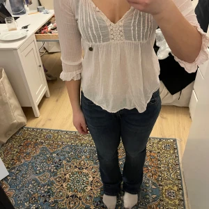 Vit blus från Noa Noa - Säljer en söt vit blus från Noa Noa med v-ringning och spetsdetaljer. Blusen har trekvartsärmar med volang vid ärmsluten och en knappdetalj framtill. Perfekt för en bohemisk look!