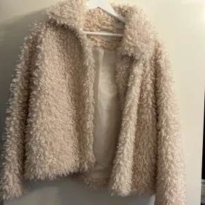 Beige teddyjacka från Kappahl - Mysig beige teddyjacka från Kappahl. Perfekt för kyliga dagar med sitt fluffiga material och stilrena design. Jackan har en klassisk krage och är helfodrad för extra värme. Ett måste för garderoben!