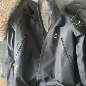 Svart vindjacka från Haglöfs - Säljer en svart vindjacka från Haglöfs med dragkedja och huva. Jackan har två framfickor med dragkedjor och är perfekt för utomhusaktiviteter. Insidan är ljusgrå och materialet är av hög kvalitet för att stå emot väder och vind.