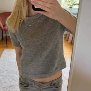 Grå stickad t-shirt från Zara - Säljer en stilren grå stickad t-shirt från Zara. Tröjan har korta ärmar och en rund halsringning, perfekt för lager på lager eller som den är. Den är mjuk och bekväm, idealisk för både vardag och avslappnade tillfällen.