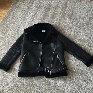 Svart skinnjacka från NA-KD - Snygg svart skinnjacka från NA-KD med dragkedja och bälte i midjan. Jackan har en cool design med långa ärmar och foder i pälsimitation. Perfekt för att ge din outfit en edgy touch.