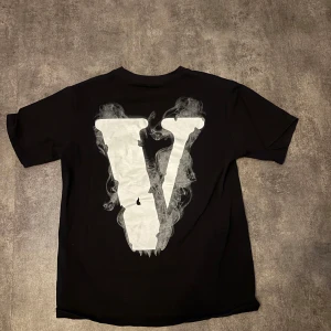 Svart t-shirt från Vlone - Säljer en svart t-shirt från Vlone med ett stort vitt 'V' på baksidan och en cool design med en ängel och texten 'VLONE' på framsidan. Perfekt för dig som gillar streetwear och unika tryck! Tröjan passas även M