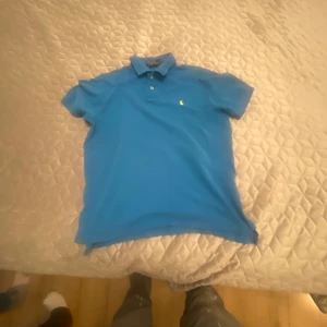 Blå pikétröja från Polo Ralph Lauren - Säljer en snygg blå pikétröja från Polo Ralph Lauren med klassisk krage och knappar. Tröjan har en gul broderad logotyp på bröstet. Perfekt för en stilren look!