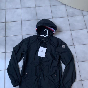 Svart vindjacka från Moncler - Säljer en snygg svart vindjacka från Moncler med huva och dragkedja. Jackan har en stilren design med Moncler-logga på ärmen och en justerbar huva med snörning. Perfekt för blåsiga dagar!