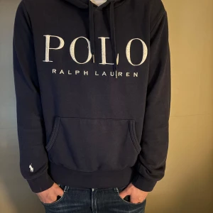 Mörkblå hoodie från Ralph Lauren - Säljer en snygg mörkblå hoodie från Ralph Lauren med stor logga på bröstet. Tröjan har en klassisk känguruficka och justerbar huva med snören. Perfekt för en avslappnad stil.