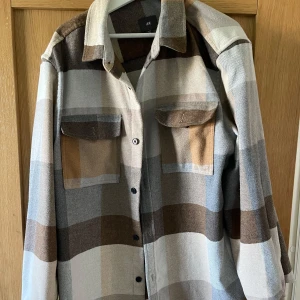 Rutig overshirt i jordnära toner - Säljer en stilren rutig overshirt i jordnära färger som beige, brun och grå. Jackan har långa ärmar och knappar framtill samt två bröstfickor med lock. Perfekt för lager-på-lager-stil under höst och vår.