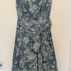 Blommig klänning med knytband - Elegant klänning i en dämpad blå nyans med vackert blommönster i vitt. Klänningen har en v-ringad hals och ett knytband i midjan som ger en smickrande passform. Perfekt för sommarens festligheter.