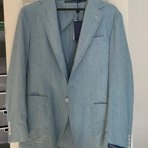 Ljusblå kavaj från Polo Ralph Lauren - Snygg ljusblå kavaj från Polo Ralph Lauren i chambray-tyg. Kavajen har en klassisk design med två knappar framtill och fyra dekorativa knappar vid ärmsluten. Perfekt för en stilren look. Ordinarie pris: 7646,54 sek