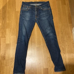 Blå jeans från Nudie Jeans - Snygga blå jeans från Nudie Jeans med klassisk femficksdesign och orange sömmar. De har en normal passform och är tillverkade i slitstarkt denim. Perfekta för en avslappnad stil. Dem har ett litet håll i fickan.