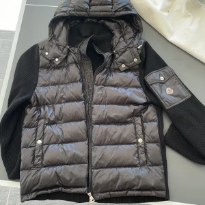 Moncler cardigan - Snygg svart dunväst från Moncler med avtagbar huva och dragkedja. Västen har stickade ärmar och en praktisk ficka på ärmen med knapp. Perfekt för kyliga dagar när du vill hålla stilen. 
