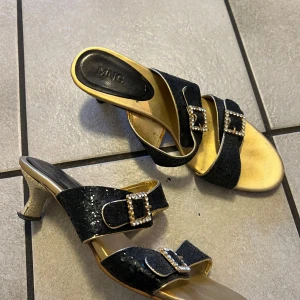Svarta och guldiga sandaler från MNG - Eleganta sandaler i svart och guld med glittriga detaljer och dekorativa spännen. Perfekta för att ge en lyxig touch till din outfit. Klackarna är stilfullt formade för extra elegans.