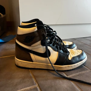 Nike Air Jordan 1 mocka  - Snygga Nike Air Jordans i svart och vit med klassisk snörning. Skorna har en hög design med det ikoniska Air Jordan-märket på sidan. Perfekta för dig som vill ha en stilren och sportig look.