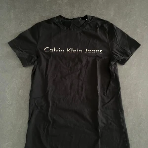 Svart t-shirt från Calvin Klein Jeans - Säljer en stilren svart t-shirt från Calvin Klein Jeans. Den är tillverkad i 100% bomull och har korta ärmar. Perfekt för en casual look med den ikoniska loggan tryckt på framsidan. Passar utmärkt till jeans eller shorts!