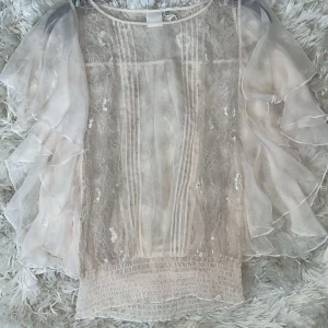 Rosa blus med volangärmar - Elegant rosa blus med volangärmar och subtila glittriga detaljer. Blusen har en luftig passform och är perfekt för en stilfull look. Den har en rund hals och är tillverkad i ett lätt och transparent material.