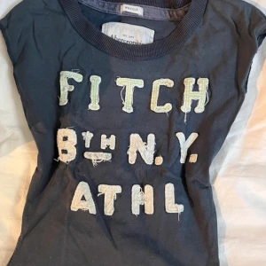 Mörkblå t-shirt från Abercrombie & Fitch - Säljer en mörkblå t-shirt från Abercrombie & Fitch med texten 'FITCH 8TH N.Y. ATHL' i slitna bokstäver på framsidan.  