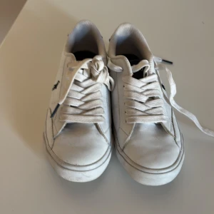 Vita sneakers från Polo - Storlek 37 Snygga vita sneakers från Polo med klassisk design och svart logga på sidan. Skorna har snörning och en rund tå, vilket ger en stilren look. Perfekta för en avslappnad stil. Dem är i bra skick men man ska vara medveten att dem är smutsiga på sullan men också snörrerna. Dem har smuts på sig men är i fint skick och använda ett par gånger 