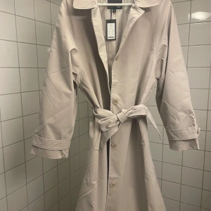 Beige trenchcoat med knytbälte - Snygg beige trenchcoat med klassisk design. Jackan har ett knytbälte i midjan och knappar framtill. Perfekt för vår och höst. Nypris 400