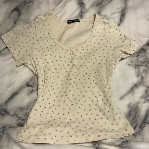 Beige blommig topp från Brandy Melville - Jättegullig beige topp med små blommor från Brandy Melville. Den har en rund halsringning med knappar framtill och korta ärmar. Jättevacker att ha på sommaren. Tröjan är använd ett fårtal gånger och har en liten fläck vid nedre delen av tröjan💕
