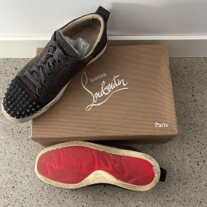 Gråa sneakers med nitar från Christian Louboutin - Snygga bruna sneakers från Christian Louboutin med nitar på tån och den ikoniska röda sulan. Skorna har snörning och är tillverkade i mocka, vilket ger en lyxig känsla. Perfekta för dig som vill sticka ut med stil. Väldigt slitna men håller någ ett bra tag till. SKRIV INNAN KÖP!🔴pris är inte hugget i sten. Box och allt ingår. 