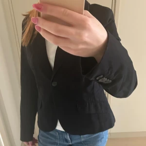 Svart kavaj från Cue - Snygg svart kavaj från Cue med broderad detalj på bröstfickan. Kavajen har en klassisk design med knappar framtill och långa ärmar. 