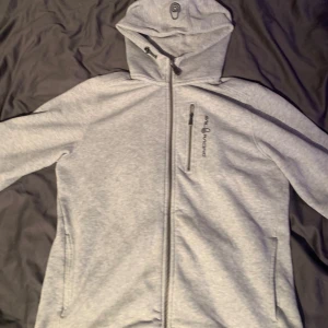 Grå hoodie från Sail Racing - Säljer en grå hoodie från Sail Racing med dragkedja och justerbar huva. Jackan har en bröstficka med dragkedja och två sidofickor. Perfekt för en avslappnad stil.