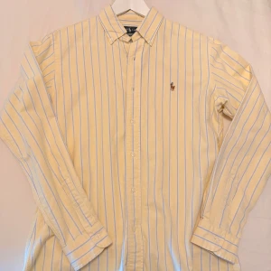 Gul och blå randig skjorta från Ralph Lauren - Säljer en stilren gul skjorta med blå ränder från Ralph Lauren. Skjortan har långa ärmar och knappar framtill. Den är broderad med den klassiska logotypen på bröstet. Perfekt för en snygg och avslappnad look. Storlek S men tycker mer den är som en M. 