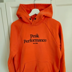 Orange hoodie från Peak Performance, S - Säljer en snygg orange hoodie från Peak Performance med svart text på bröstet. Den har en klassisk design med huva och känguruficka framtill. Perfekt för en avslappnad stil.