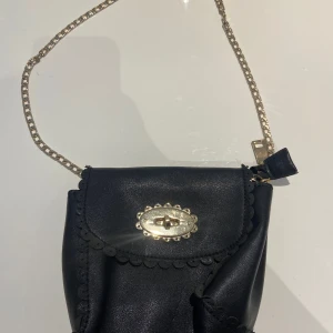 Svart axelväska med guldkedja - Snygg svart axelväska med guldkedja och dekorativt spänne. Väskan har en elegant design med volangdetaljer och en praktisk storlek för att bära dina nödvändigheter. Perfekt för att ge en stilfull touch till din outfit.