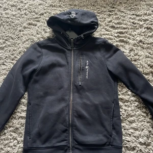 Svart hoodie från Sail Racing - Säljer en svart hoodie från Sail Racing med dragkedja och huva. Tröjan har en bröstficka med dragkedja och två sidofickor. Perfekt för kyliga dagar och en stilren look.