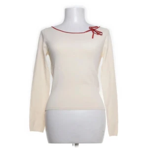 Beige tröja med rosett från Hallhuber - Säljer en stilren beige tröja från Hallhuber Essentials. Tröjan har en söt röd rosett vid halsringningen och långa ärmar. Perfekt för en elegant look. Materialet är en blandning av rayon och nylon.