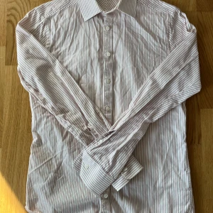 Randig skjorta från Tiger of Sweden - Säljer en stilren randig skjorta från Tiger of Sweden i vitt och grönt . Skjortan har långa ärmar och knappar framtill. Perfekt för en elegant look.