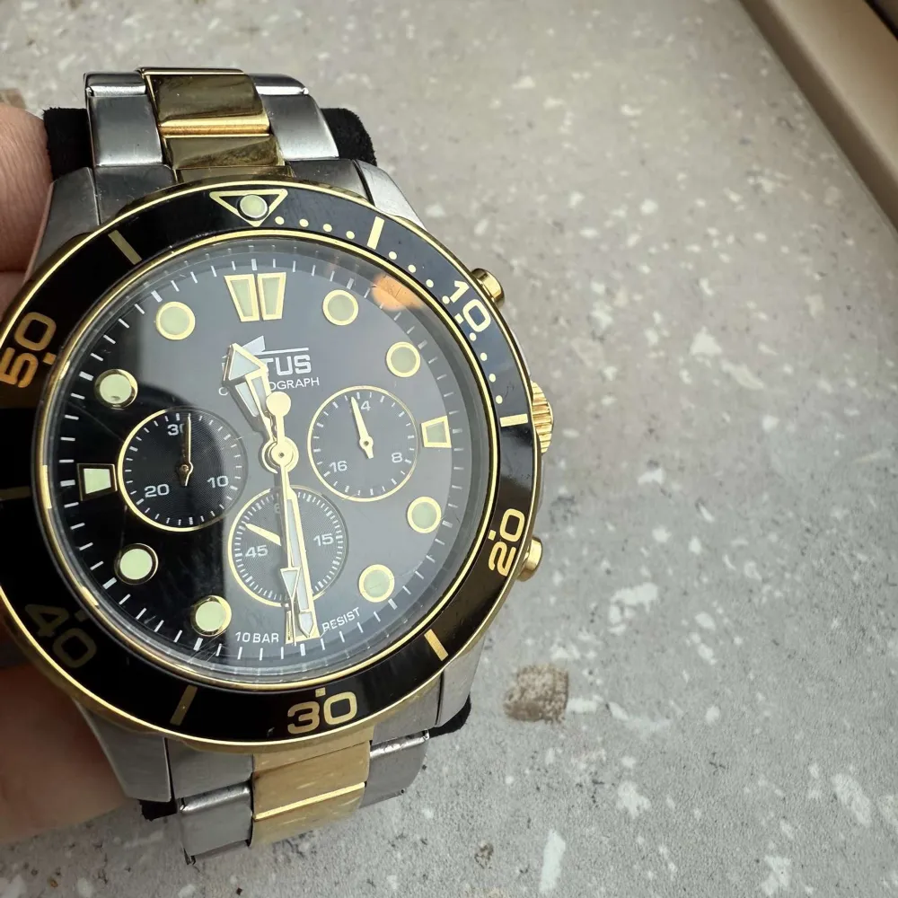 Snygg Lotus Chronograph herrklocka med en kombination av silver och guld på armbandet. Klockan har en svart urtavla med guldiga detaljer och flera sub-dials. Perfekt för den som vill ha en stilren och funktionell accessoar.. Asusteet.