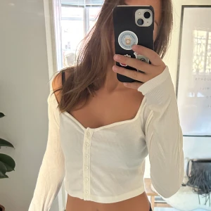 Vit ribbad offshoulder topp - Säljer en stilren vit ribbad offshoulder topp med knappar framtill. Toppen är croppad och har långa ärmar, perfekt för en trendig look. Passar bra till både jeans och kjol. Hör av dig vid intresse!