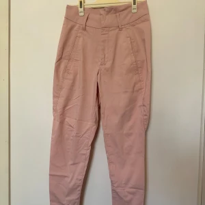 Rosa byxor från Kappahl - Rosa stretchiga byxor från Kappahl med normal passform. Byxorna har en klassisk design med bälteshällor och dragkedja.