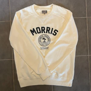 Morris Tröja Naturvit M - Säljer en stilren vit sweatshirt från Morris med svart text och ett tryck på framsidan. Tröjan har långa ärmar och en rund halsringning. Perfekt för en avslappnad stil.
