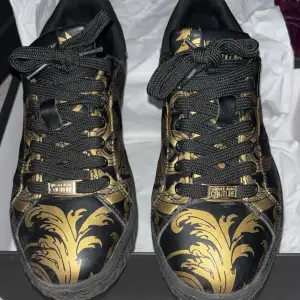 Snygga sneakers i svart med guldigt barockmönster från Versace Jeans Couture. Skorna har snörning och en rund tå, med märkets logga på sidan och hälen. 42-43 storlek. Äkta!!!.