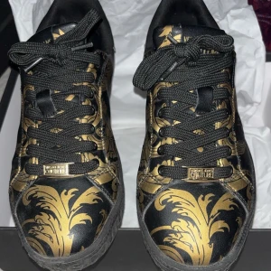 Versace skor  - Snygga sneakers i svart med guldigt barockmönster från Versace Jeans Couture. Skorna har snörning och en rund tå, med märkets logga på sidan och hälen. 42-43 storlek. Äkta!!!.