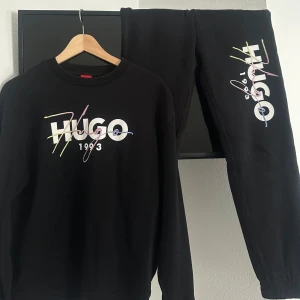 Svart sweatshirt och byxor från Hugo - Säljer en svart sweatshirt och matchande byxor från Hugo med färgglatt tryck av märkesnamnet och årtalet 1993. Tröjan har långa ärmar och en rund halsringning. Perfekt för en avslappnad stil.