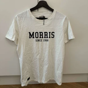 Vit t-shirt från Morris - Säljer en stilren vit t-shirt från Morris med svart text 'Morris Since 1968' på bröstet. Perfekt för en avslappnad look. T-shirten har en klassisk rund hals och korta ärmar.