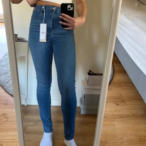 Blå högmidjade jeans - Säljer ett par snygga helt nya blå högmidjade Molly jeans med tight passform. Perfekta för en stilren look. De har en klassisk design med dragkedja och knapp framtill. Prislappen är kvar