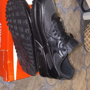Svarta Nike Air Max 90 - Säljer ett par svarta Nike Air Max med snygg design och bekväm passform. Skorna har en klassisk siluett med snörning och en robust sula för extra komfort. Perfekta för både vardag och träning.