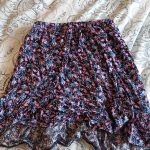 Blommig kjol från Mywear - Säljer en söt blommig kjol från Mywear i blått, rosa och svart. Kjolen har volangdetaljer och är tillverkad i mjuk viskos. Perfekt för dig som gillar färgglada och lekfulla plagg.