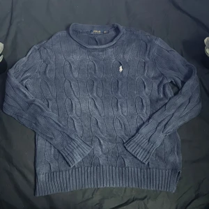 Blå kabelstickad tröja från Polo Ralph Lauren - Säljer en klassisk blå kabelstickad tröja från Polo Ralph Lauren med rund halsringning och broderad logga på bröstet. Tröjan har långa ärmar och ribbade muddar vid ärmslut och nederkant. Perfekt för en stilren look.